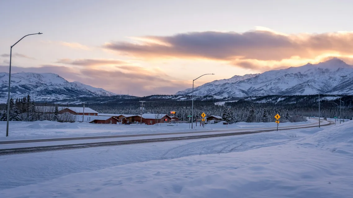 A quiet snowy road in Fairbanks, Alaska, stretching towards a cluster of buildings with a backdrop of snow-covered mountains and a glowing sunset sky. The scene captures the calm and beauty of winter in a remote northern town.