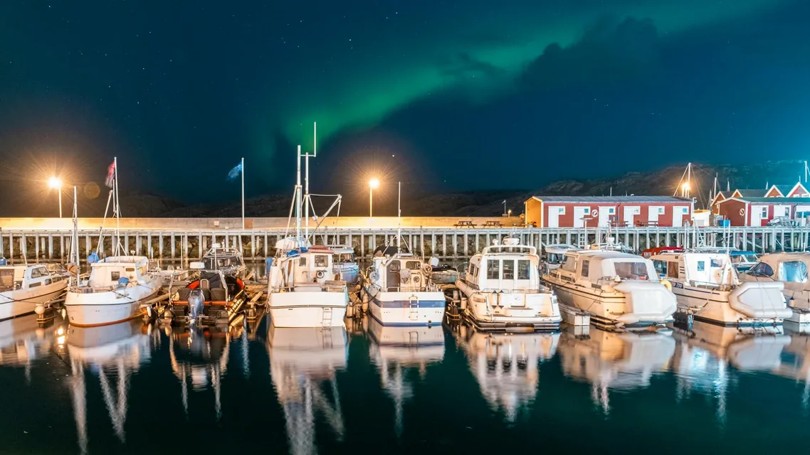 A row of white boats docked in the harbour at Bodø, as an aurora lights up the sky above